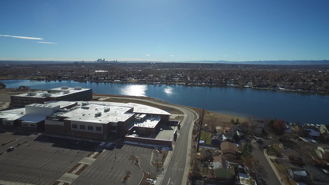 Aerial frame in Westminster, Colorado with the building roof centered, mostly empty parking in front, bright sun reflection on the river, and no visible people.