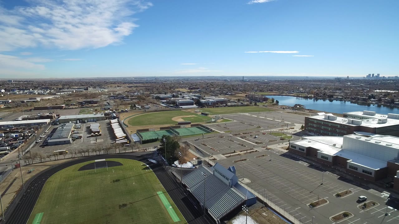 Aerial view of a football field, track, bleachers, parking lots, lake, and distant skyline.