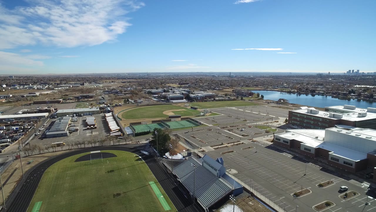 Aerial frame in Westminster, Colorado with the field and black track in the foreground, bleachers at lower right, broad parking areas at center, and no visible people.