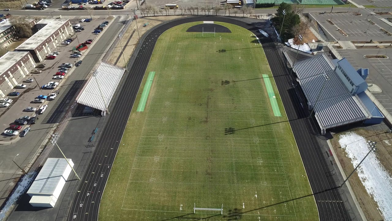 Aerial frame over a Westminster, Colorado sports field with a black running track, white bleachers, nearby parked cars, and tiny distant people on the turf.