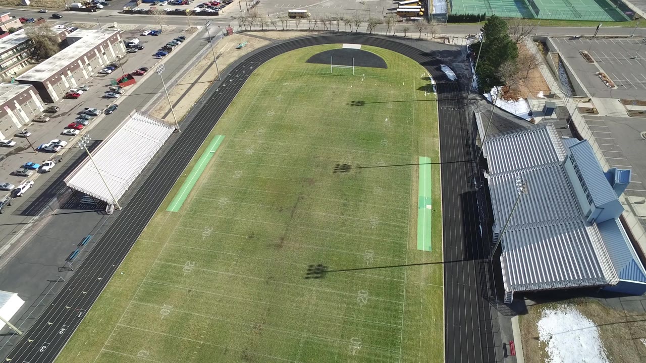 Drone perspective of the Westminster, Colorado stadium showing the football field, end zone track curve, metal bleachers, parking rows, and no visible people.