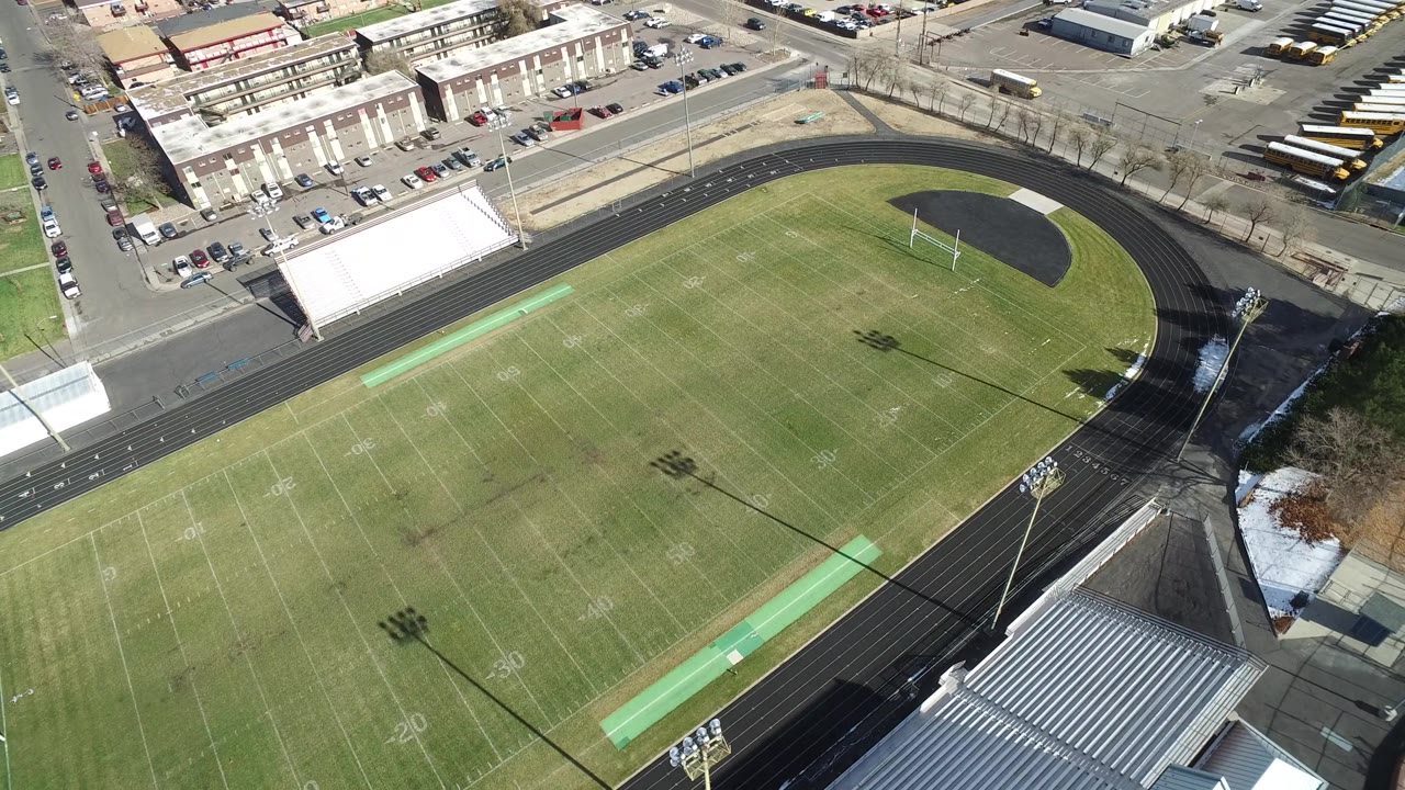 High aerial frame of the Westminster football stadium, showing yard lines, goalposts, a curved track, empty stands, nearby buildings, and no visible people.