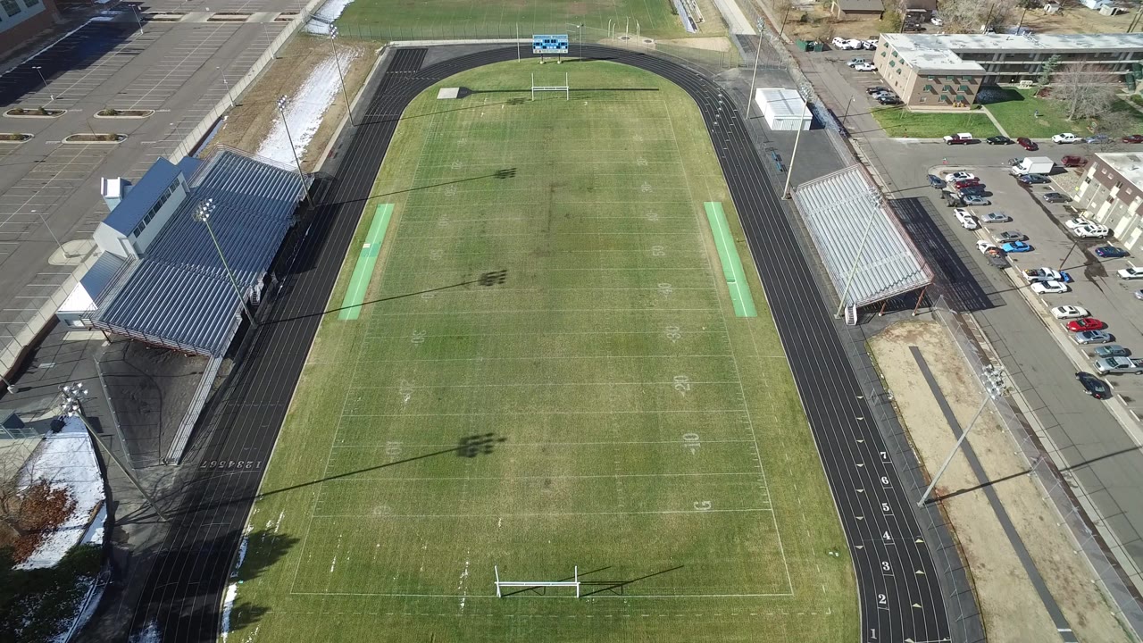 Top-down frame of the Westminster, Colorado sports field with yard lines, goalposts, a black oval track, metal bleachers, parked cars, and no visible people.