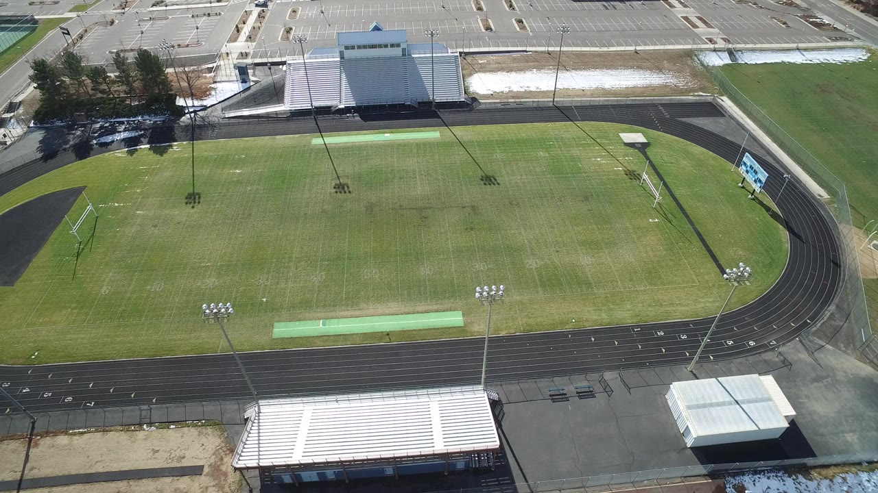 Aerial view over an empty Westminster, Colorado football stadium with a green field, black running track, white bleachers, parking lot, and no visible people.