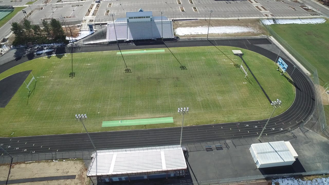 High aerial frame of the Westminster stadium showing more of the field center, the curved black track, metal bleachers, parking spaces, and no visible people.