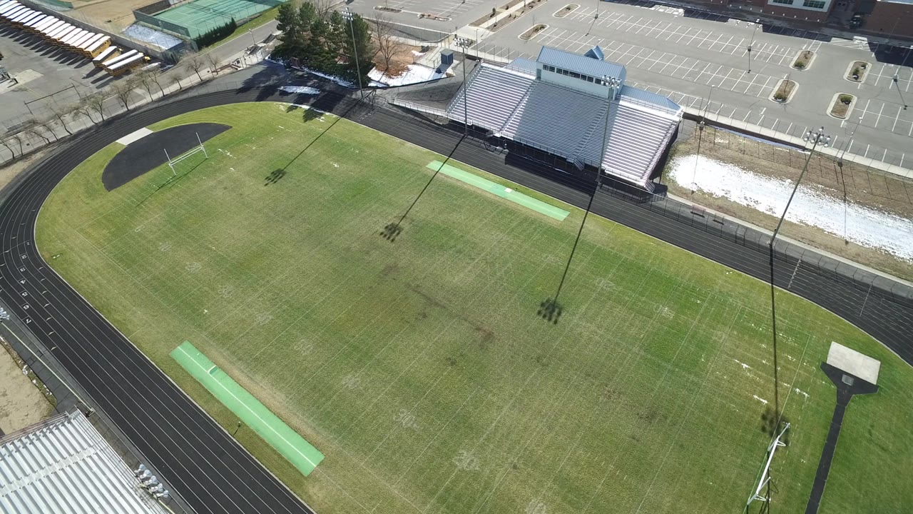 A slightly shifted aerial frame in Westminster, Colorado shows the curved track around the field, metal bleachers at right, an empty parking lot, long pole shadows, and no visible people.