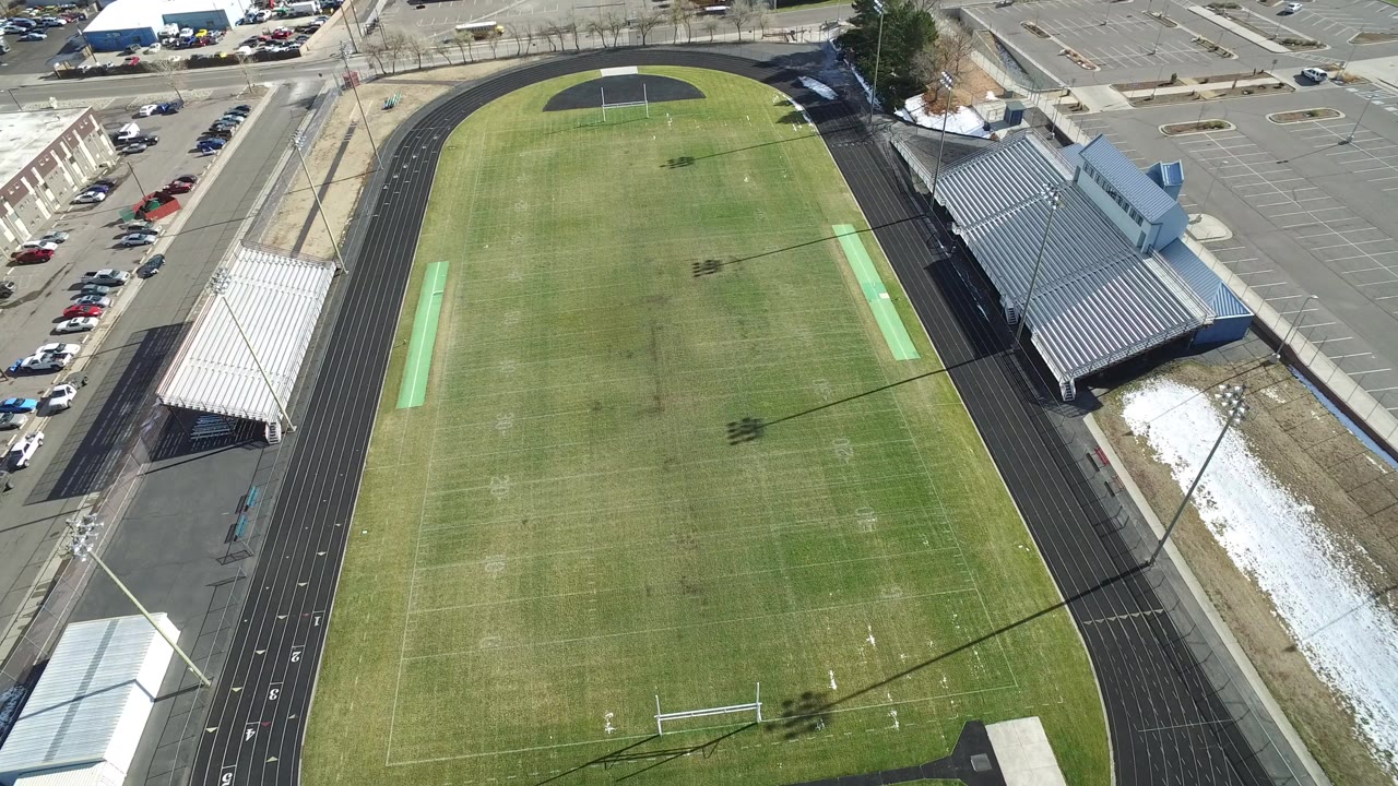 High aerial frame over a Westminster, Colorado football field inside a black running track, with bleachers, parked cars, streets, snow patches, and no visible people.