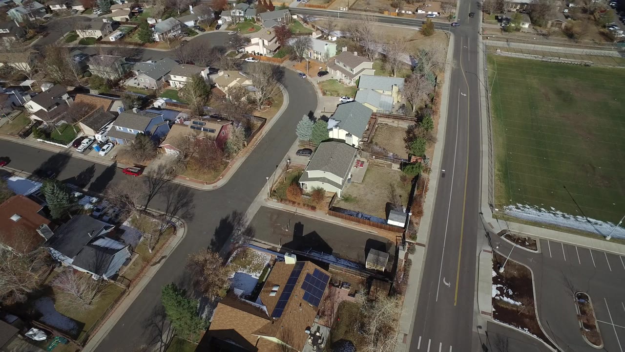 Overhead view of Westminster, Colorado rooftops, fenced yards, parked vehicles, curving streets, a field, and no visible people.