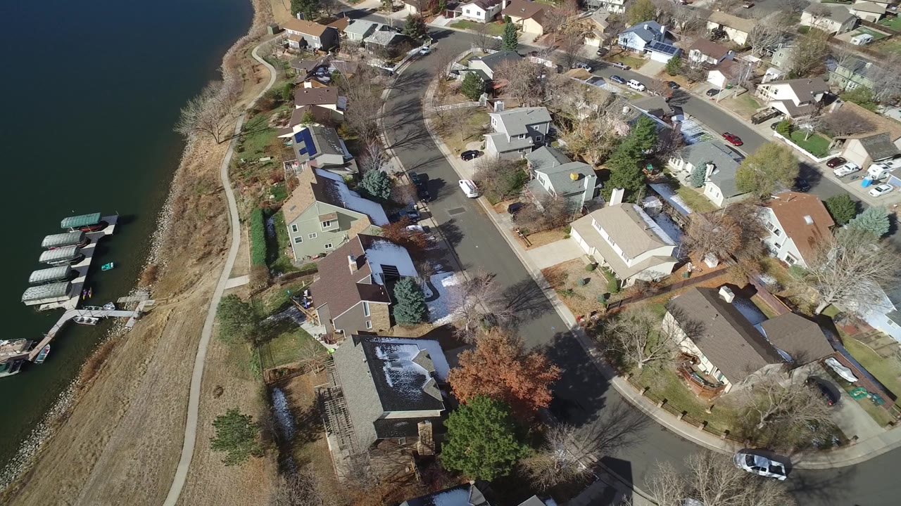Aerial frame in Westminster, Colorado showing dark lake water beside a strip of shoreline, covered boat slips, detached houses, a curved street, parked cars, and no visible people.