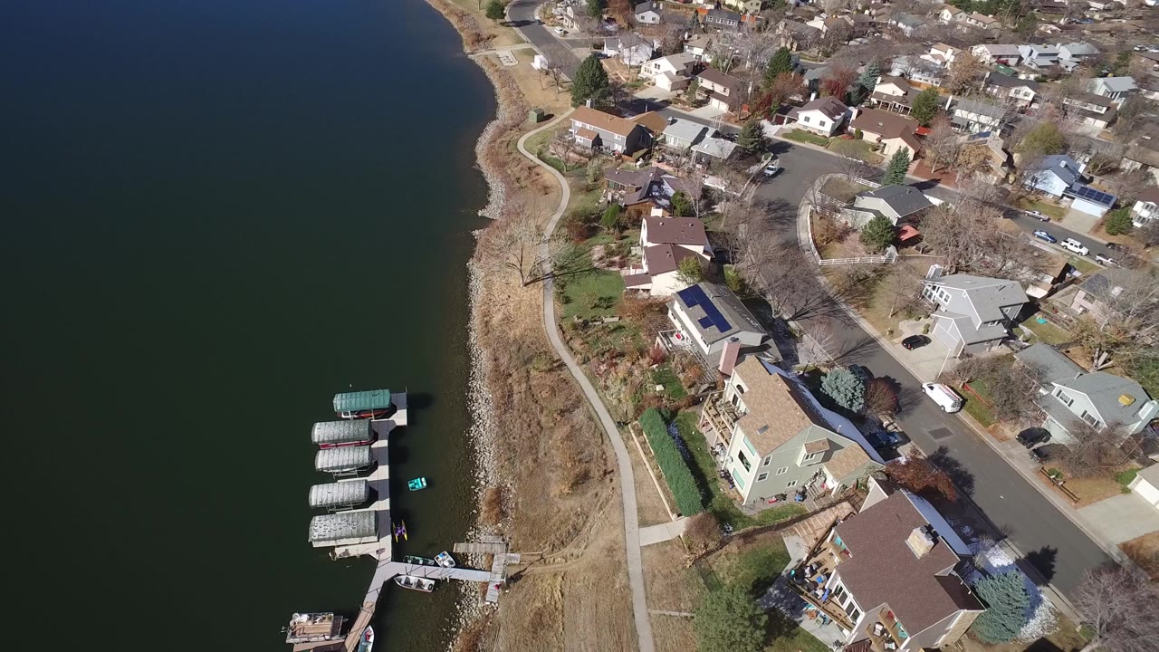 Aerial view in Westminster, Colorado with lake water on the left, a small marina-like dock area near the lower edge, residential roofs, roads, parked vehicles, and no visible people.