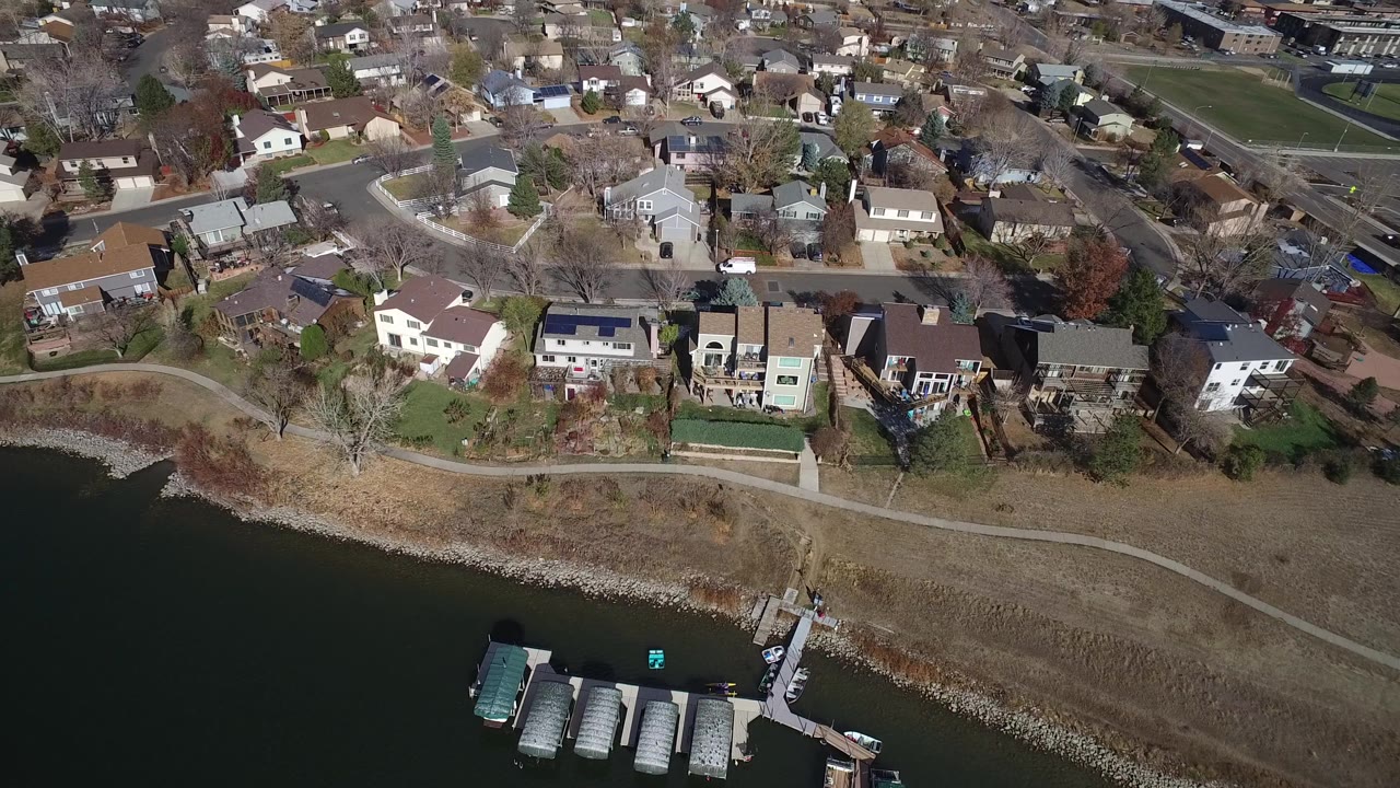 High aerial view over Westminster, Colorado homes along a shoreline with docks, boats, roads, dry grass, leafless trees, and no visible people.