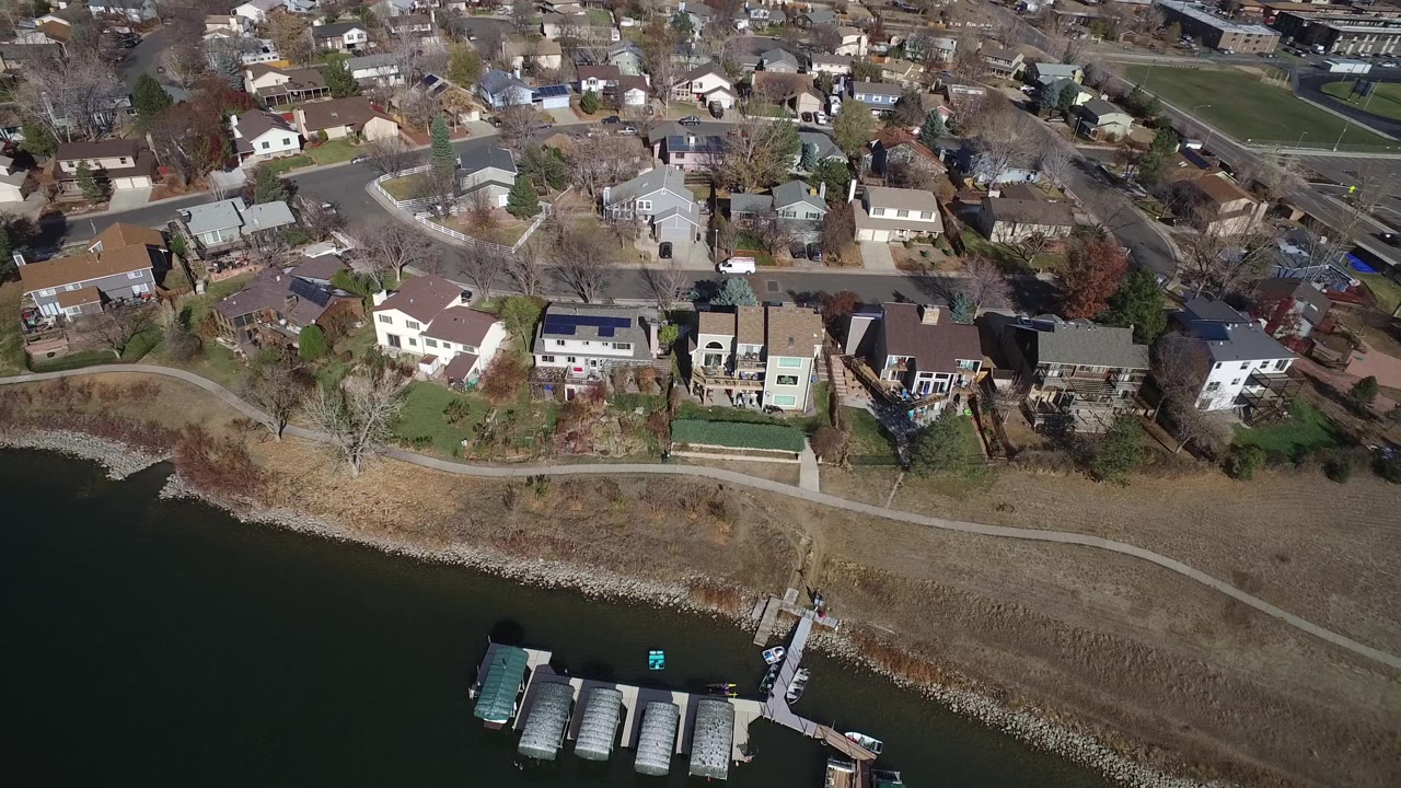 Aerial frame in Westminster, Colorado showing waterfront homes, several docks with boats, a curved footpath, residential streets, winter-brown grass, and no visible people.
