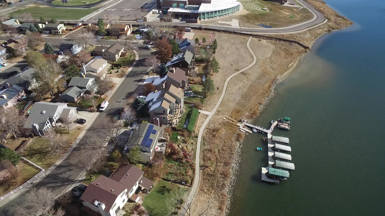High aerial frame over Westminster, Colorado showing lakeside houses, a curved shore path, docks with covered boats, open water, and no visible people.