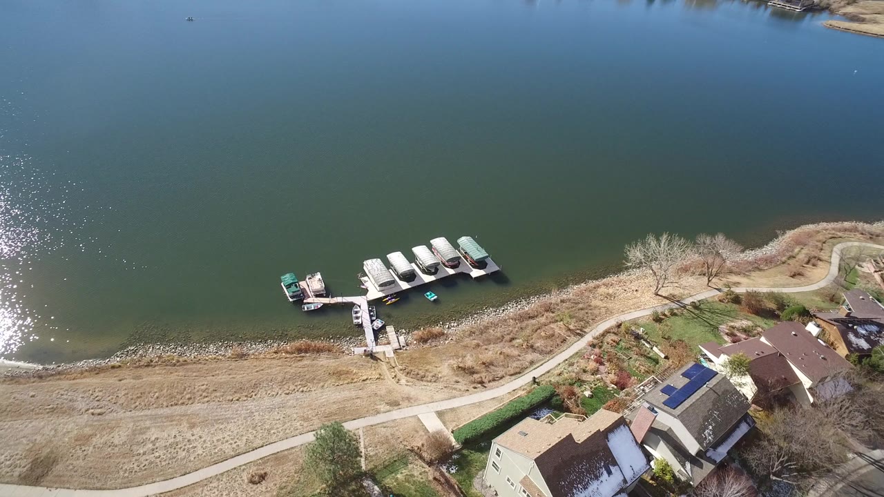 High aerial frame of Westminster, Colorado shoreline where covered boats and a narrow dock sit below houses with roofs and yards, with no visible people.