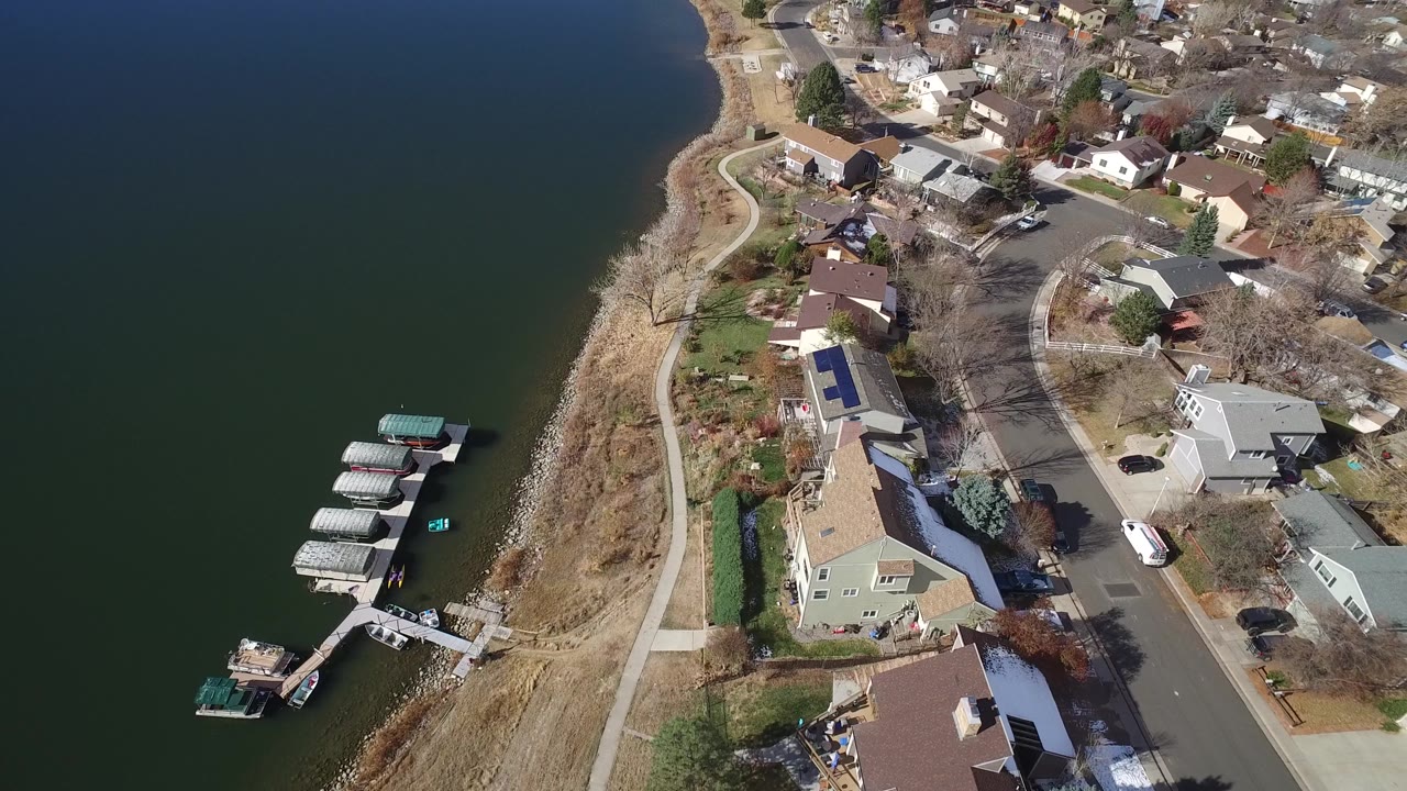 Aerial frame in Westminster, Colorado with a covered dock cluster near the lower left, lakeside houses, a road, bare trees, and no visible people.