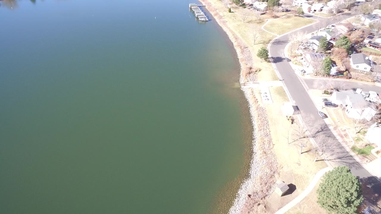 Aerial frame in Westminster, Colorado showing the lake filling most of the view, a narrow rocky bank, nearby houses, lawns, and a curved neighborhood road, no visible people.