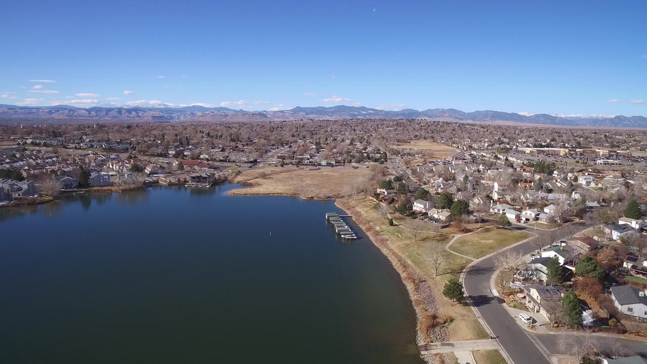 High aerial view in Westminster, Colorado looking across a dark lake toward shoreline homes, a curved road, small docks, and distant mountains under clear sky, with no visible people.