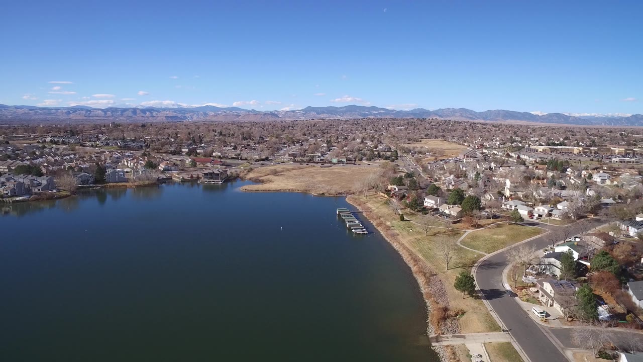 Wide aerial over a suburban lake with docks, homes, roads, and mountains.
