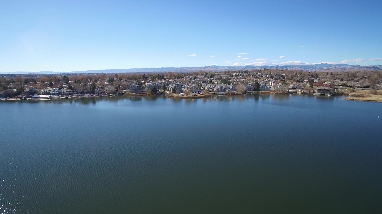Wide lake with distant houses, trees, mountains, and blue sky.
