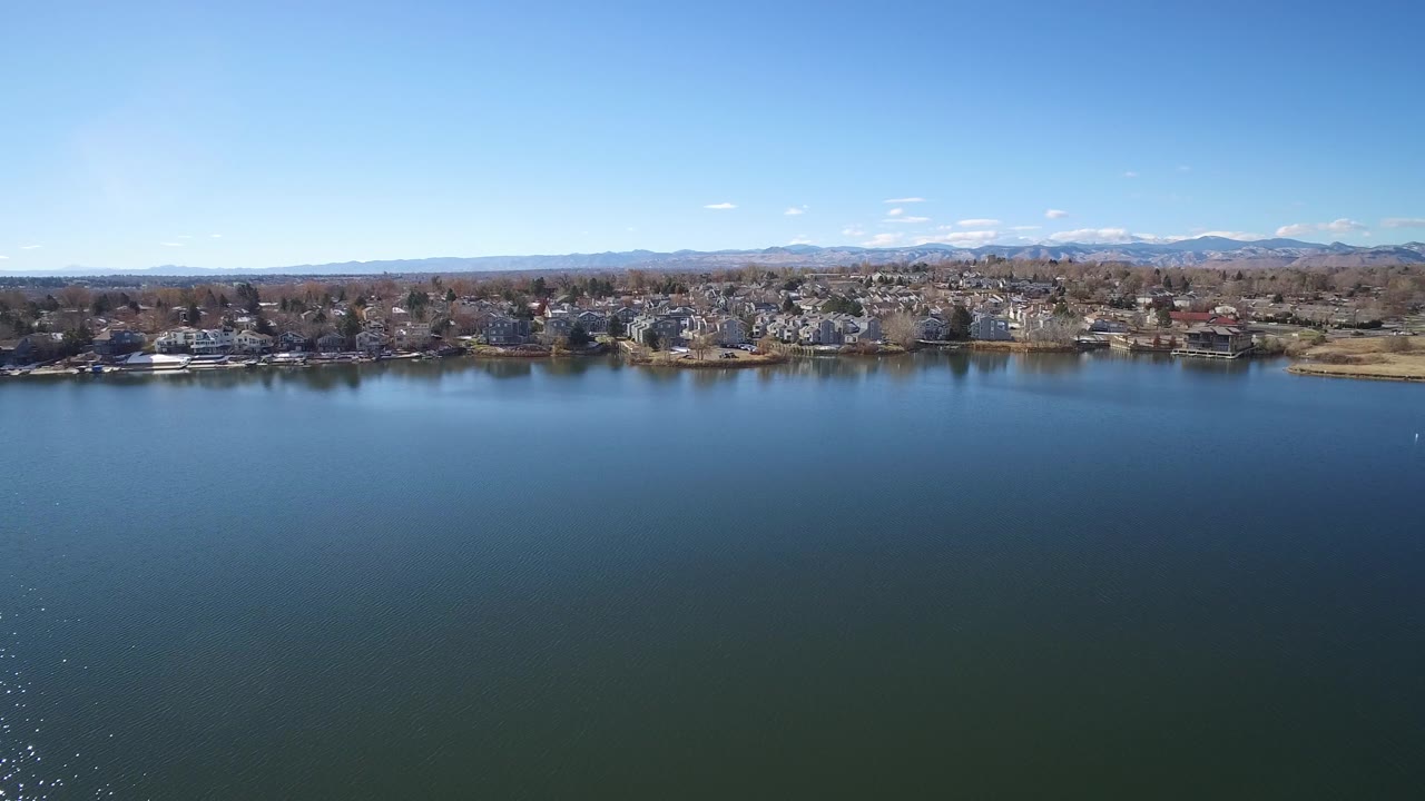A broad Westminster, Colorado lake view shows still blue water, a band of houses and trees along the far shore, low mountains beyond, and no visible people.