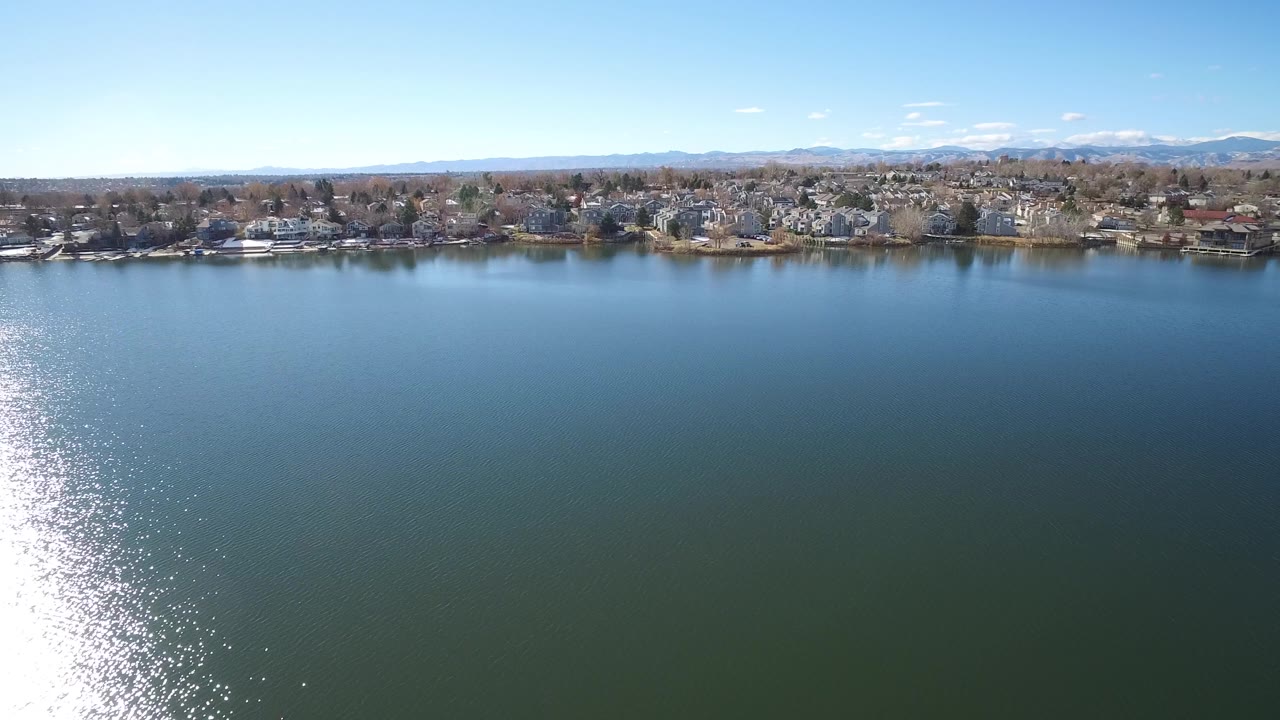 Aerial lake view in Westminster, Colorado, with the shoreline centered across the frame, houses partly screened by leafless trees, distant mountains, and no visible people.