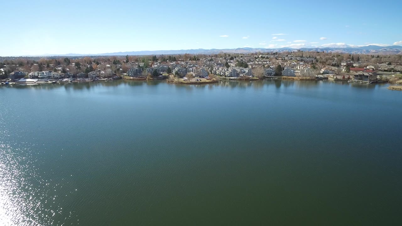 Aerial view across lake water toward suburban shoreline and distant mountains.