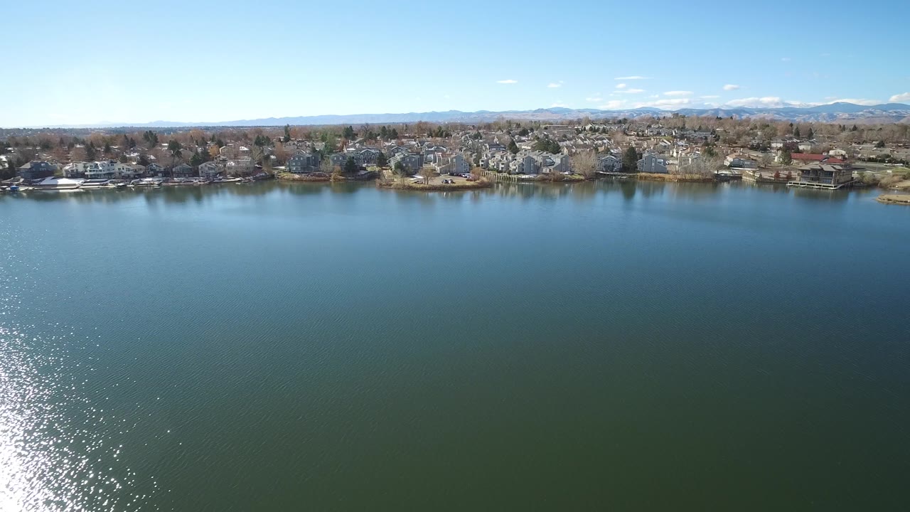 Westminster, Colorado aerial view with calm lake water filling most of the frame, small shoreline buildings in the distance, a bright water reflection, mountain ridge, clear sky, and no visible people.