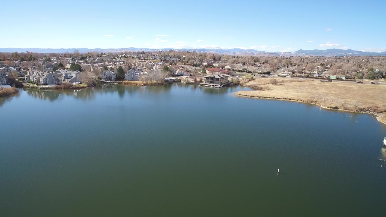 Aerial view over greenish lake water with homes, shoreline, and distant mountains.