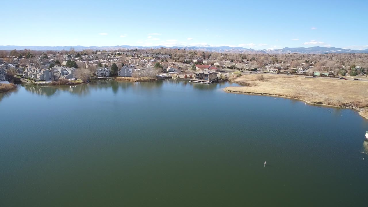Aerial view in Westminster, Colorado, looking across greenish water toward houses and leafless trees, with the dry right shoreline more centered and no visible people.