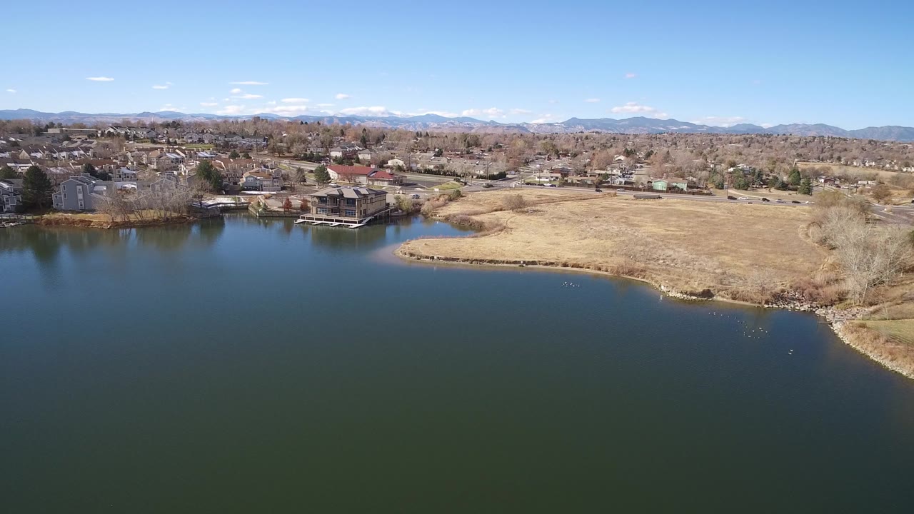 Aerial view in Westminster, Colorado looking over dark lake water toward a tan shoreline, lakefront homes, leafless trees, and distant mountains under a clear sky, with no visible people.