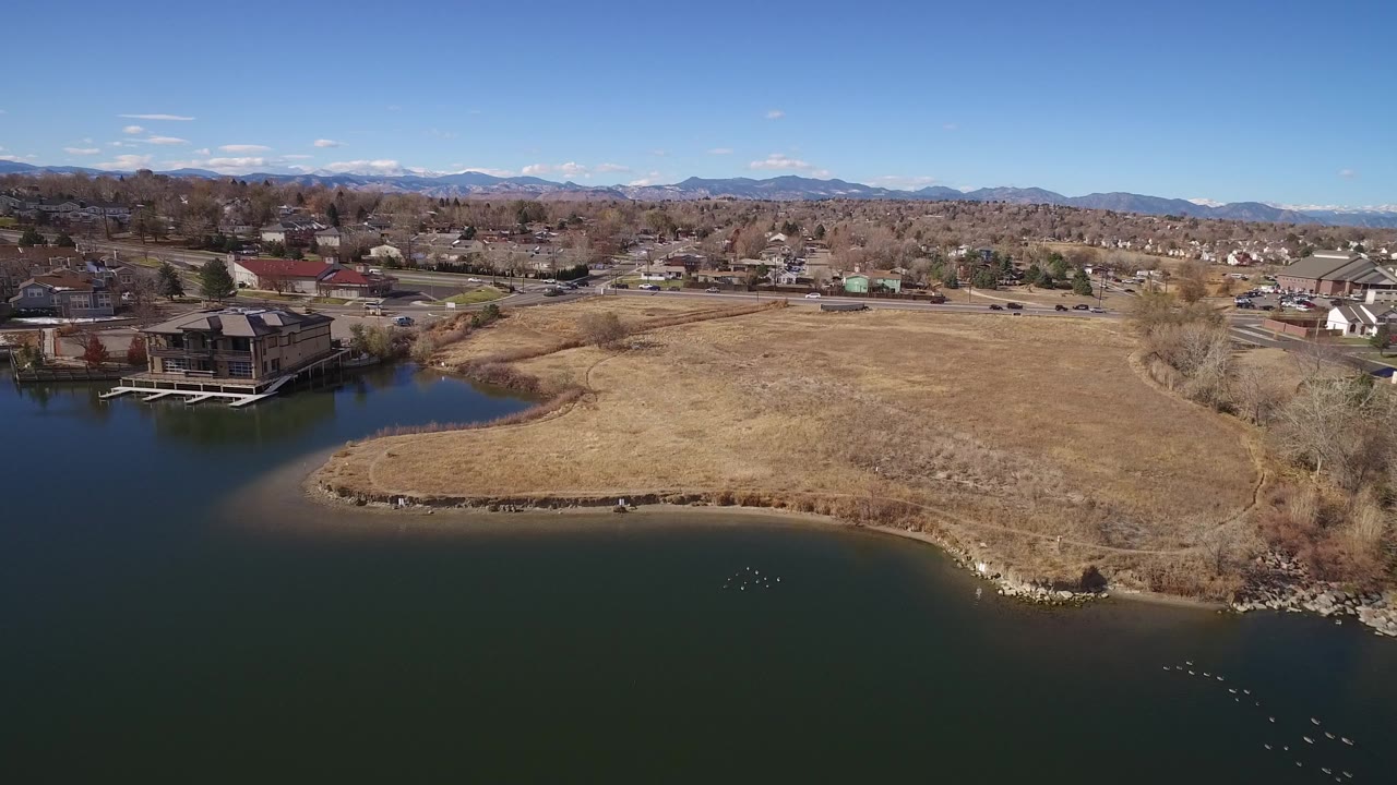 Aerial view of pond shoreline, dry grass, homes, trees, and distant mountains.