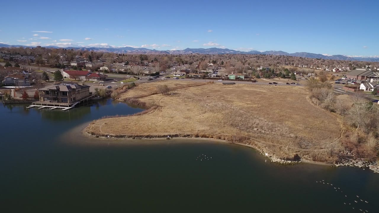 Aerial view in Westminster, Colorado looking across pond water and a curved tan shoreline toward open grass, houses, leafless trees, roads, mountains, blue sky, and no visible people.
