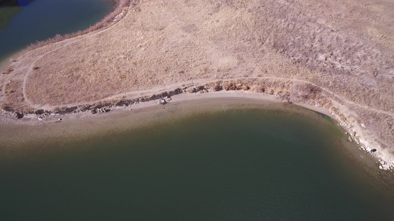Overhead aerial frame from Westminster, Colorado shows dark water along a curved sandy beach with tan dry grass beyond the shore and no visible people.