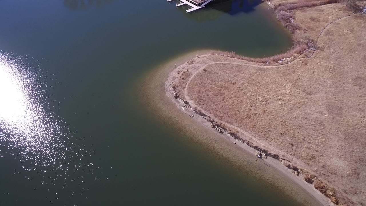 Aerial frame over Westminster, Colorado water with a dry shoreline point centered lower in frame and bright reflected sun along the left edge, with no visible people.