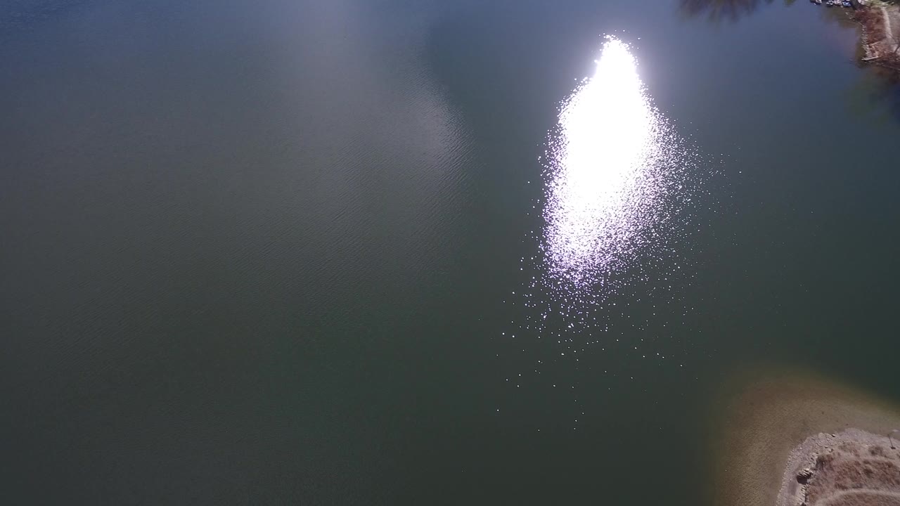 Downward aerial view of dark lake water with bright sun reflection.