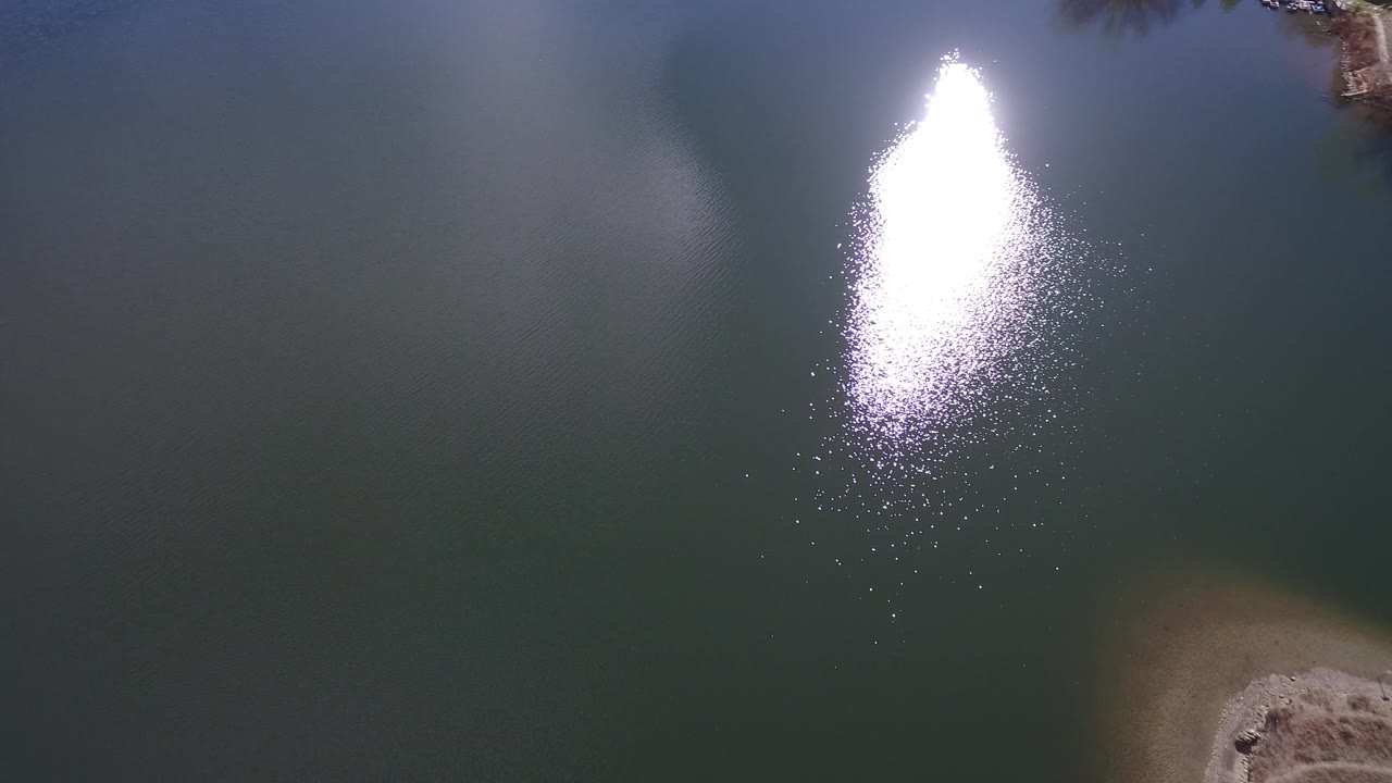 Aerial view of blue-green Westminster lake water with the sun glare shifted slightly right and a small shoreline detail near the lower edge, with no visible people.