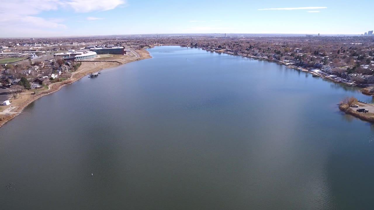 Aerial view in Westminster, Colorado over a wide blue-green waterway, with tan shoreline, houses, low buildings, and a distant skyline under a pale sky, no visible people.