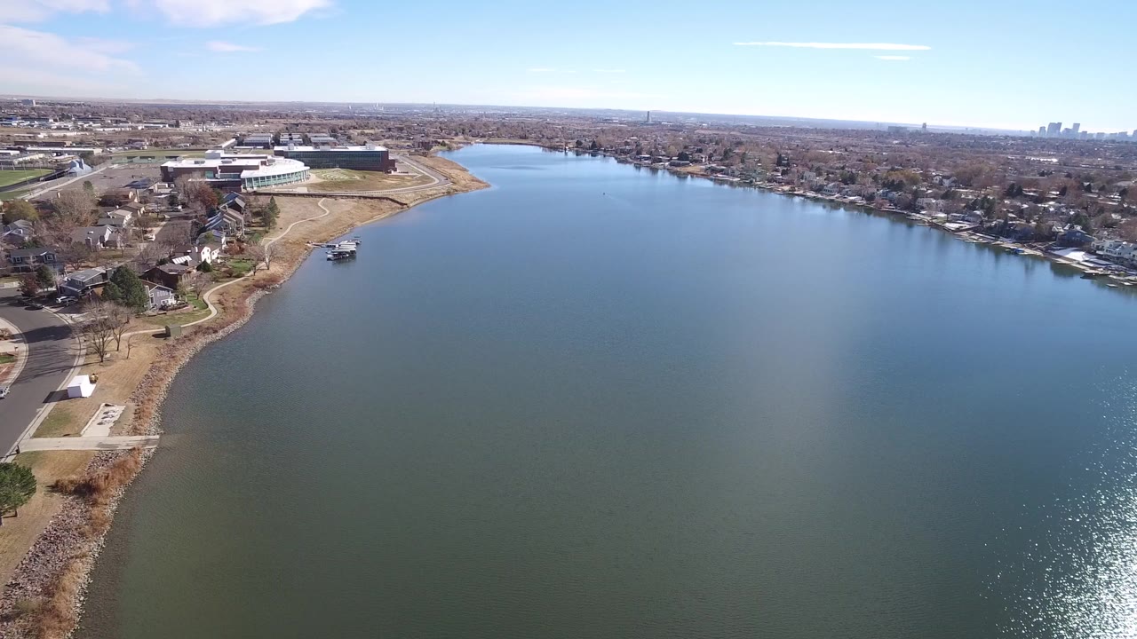 Aerial frame over water in Westminster, Colorado, with a shoreline road, houses, bare trees, low buildings, distant skyline, bright water glare, and no visible people.