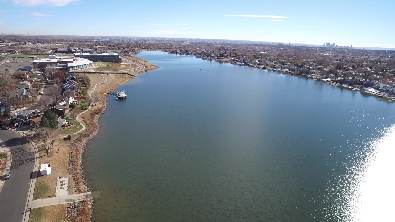 High view of a lake shoreline with houses, a road, large buildings, and bright reflection on the water.