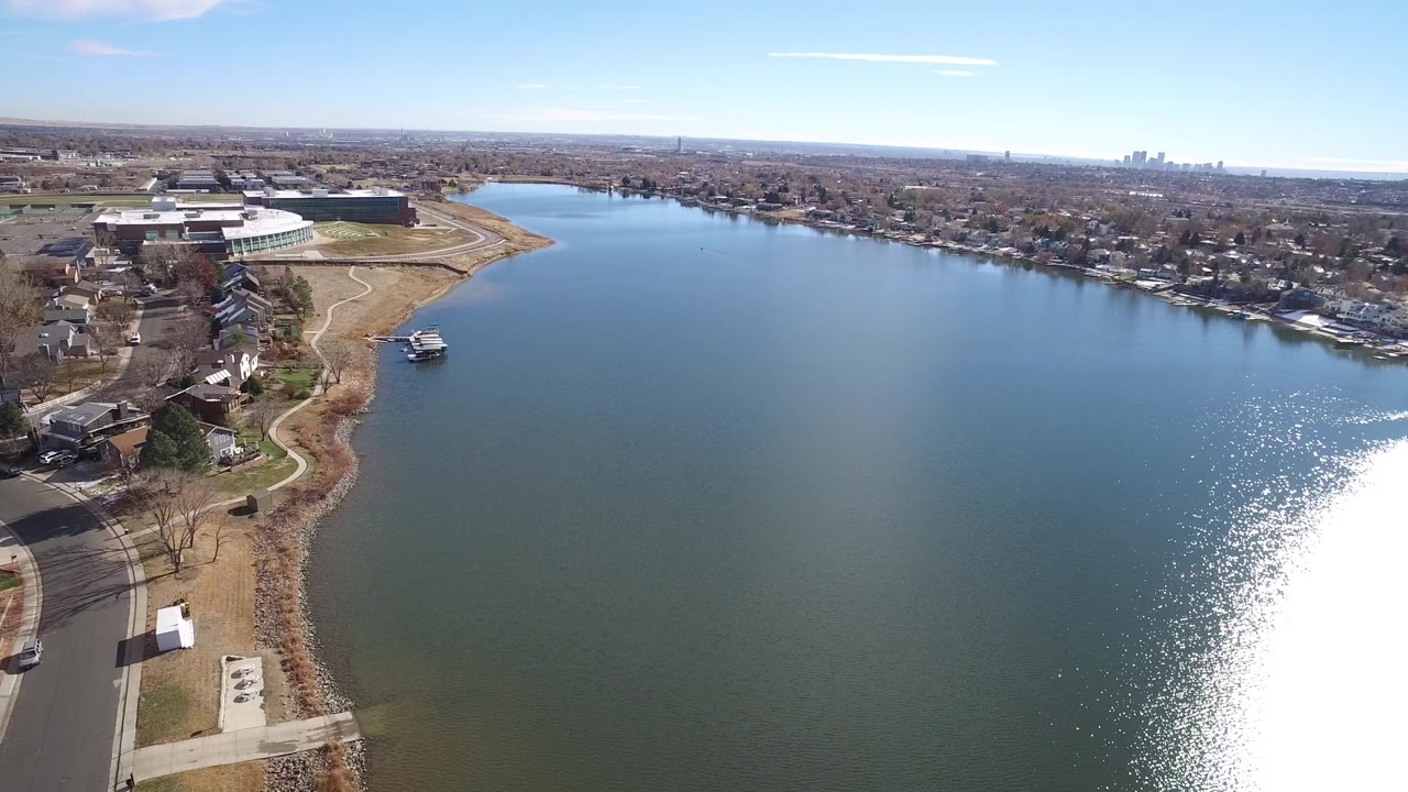High aerial view in Westminster, Colorado with the lake filling most of the frame, homes and a curved road along the near shore, large buildings on the far left, distant skyline, and no visible people.