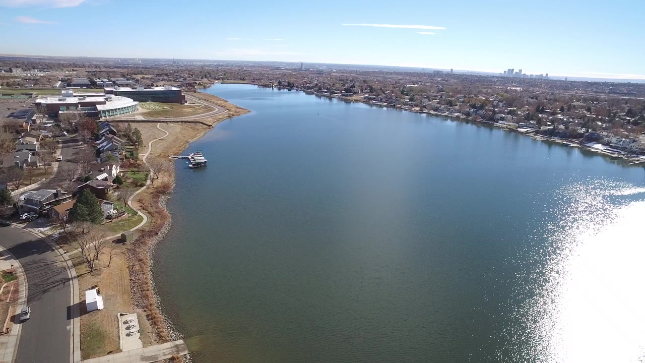 Aerial view across a lake with shoreline homes, tan grass, paths, and strong sun sparkle.