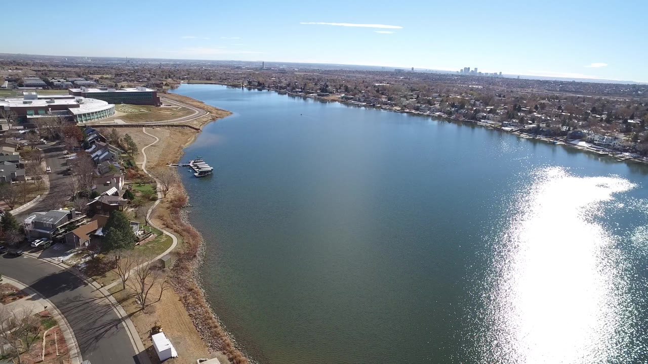 Aerial view over broad water, shoreline homes, paths, large buildings, and distant skyline.