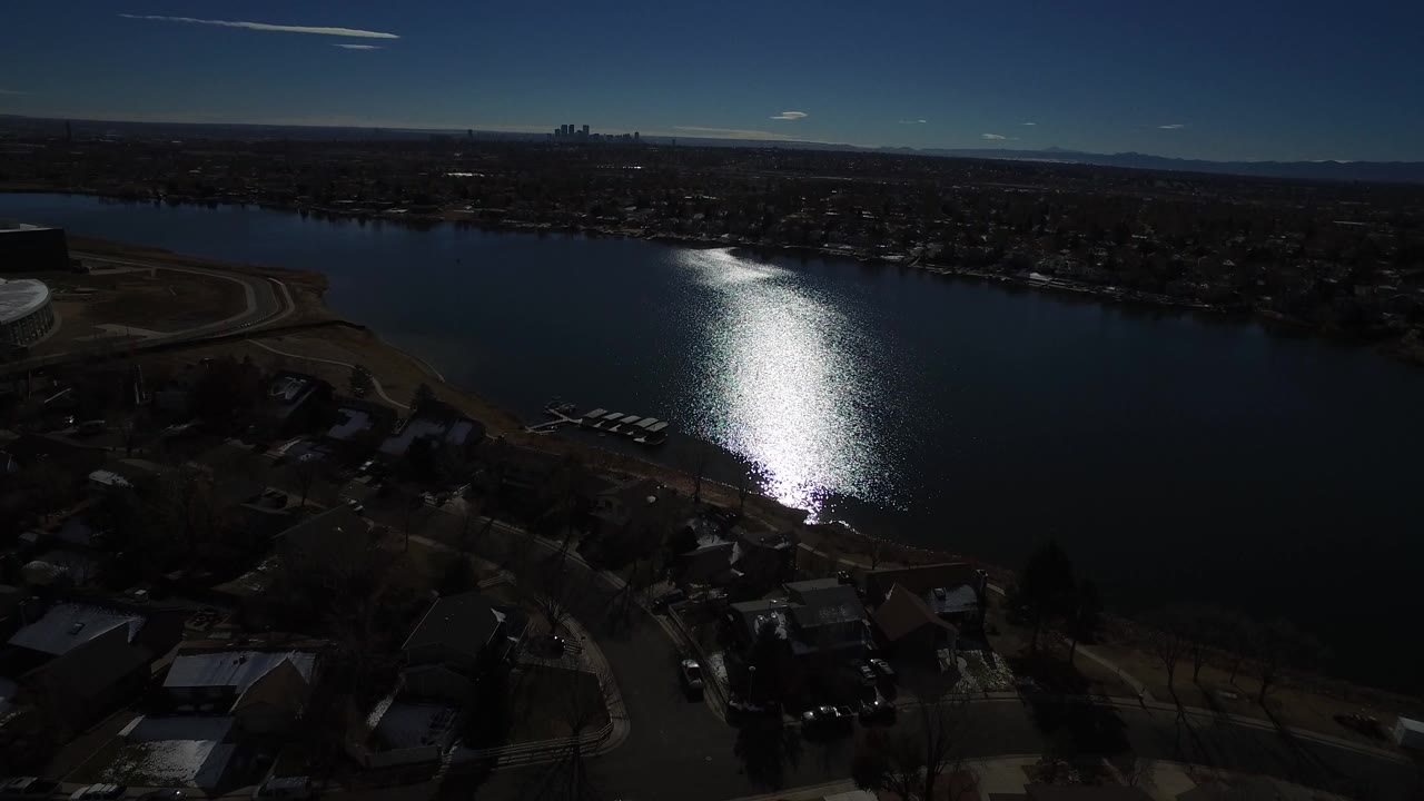 Aerial view of a lake with sun glare, homes, roads, and distant skyline.