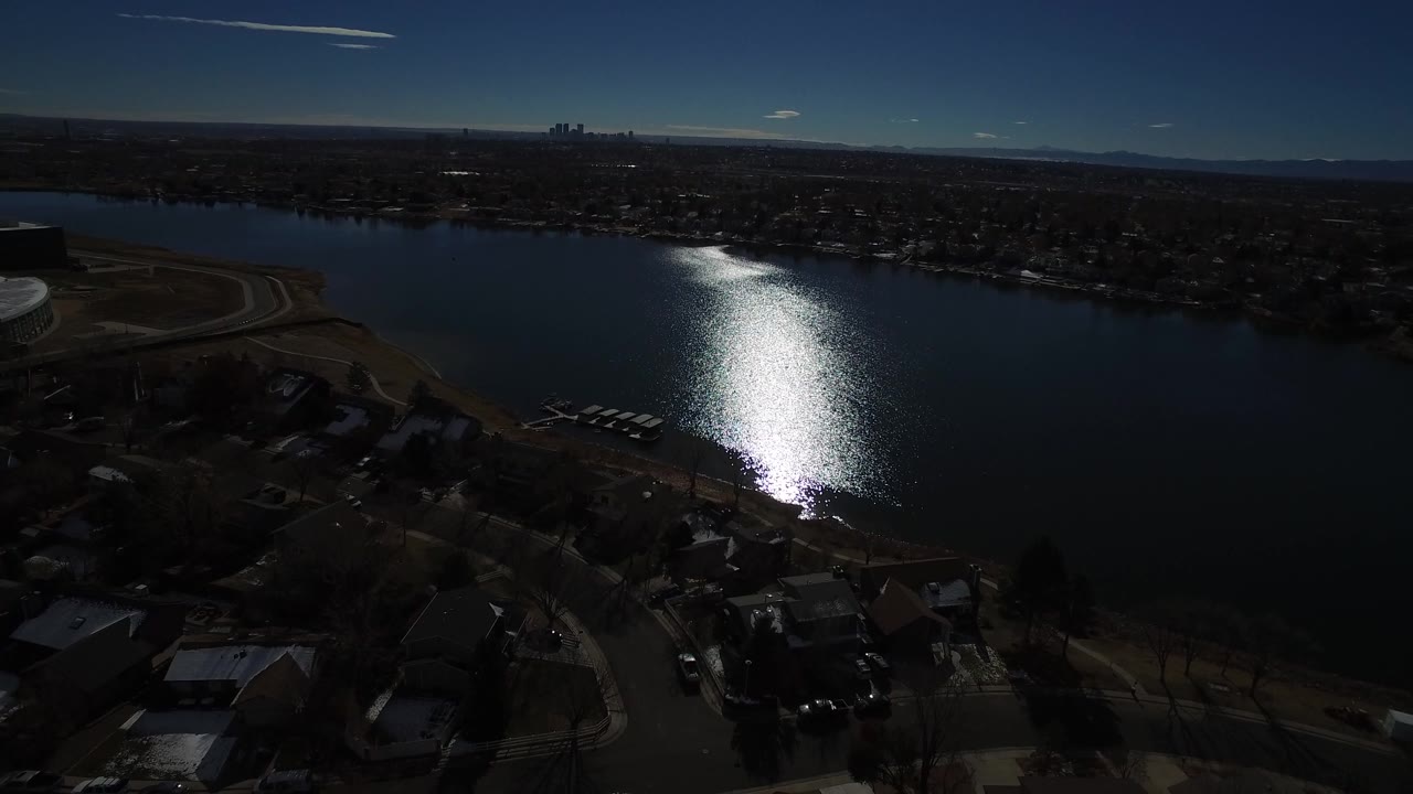 Westminster, Colorado aerial view with the sun glare centered on the lake, residential roofs and roads in shadow below, skyline on the far shore, and no visible people.
