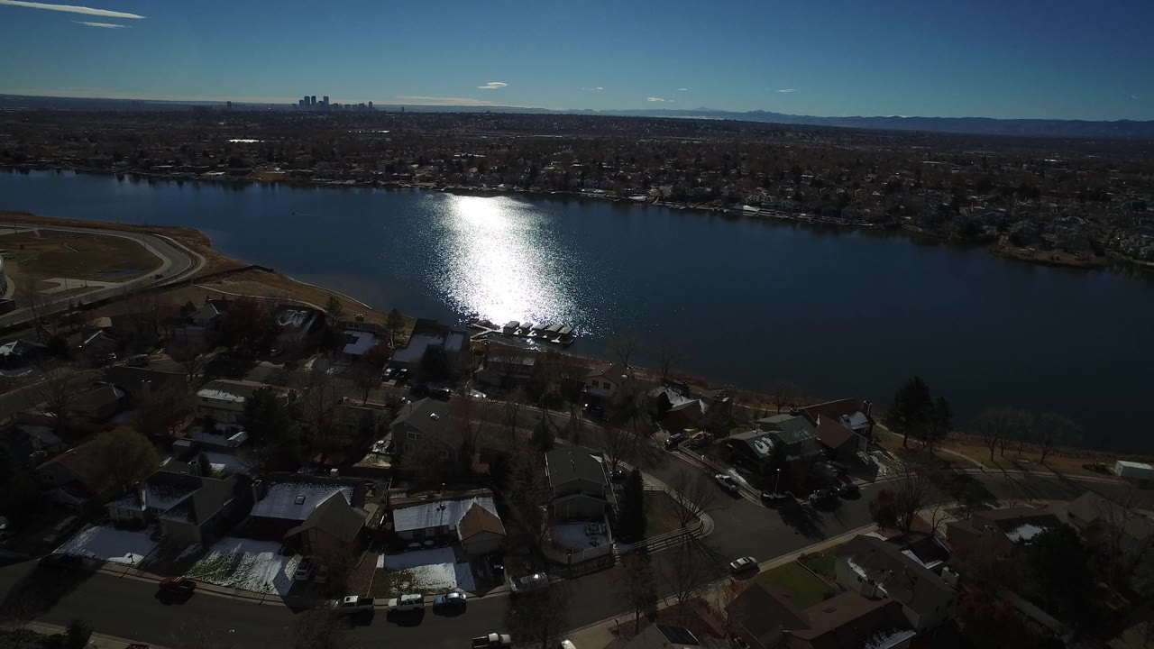 High aerial view in Westminster, Colorado with foreground rooftops and streets, a large blue lake, bright water glare near the shore, distant buildings, and no visible people.