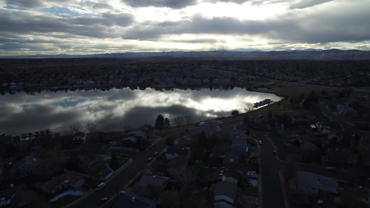 Aerial view over dark suburban homes toward a reflective lake and cloudy horizon.
