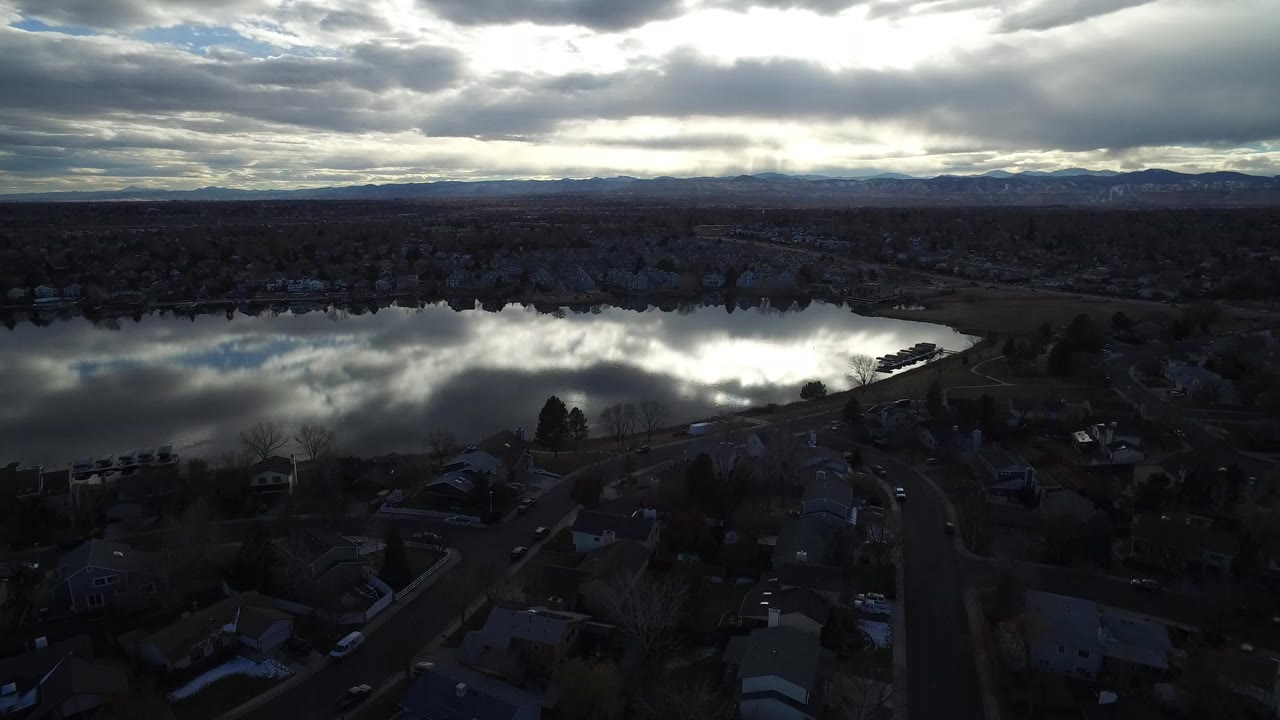 Westminster, Colorado aerial view with the lake centered across the frame, dark homes and trees below, distant hills on the horizon, heavy dusk clouds, and no visible people.