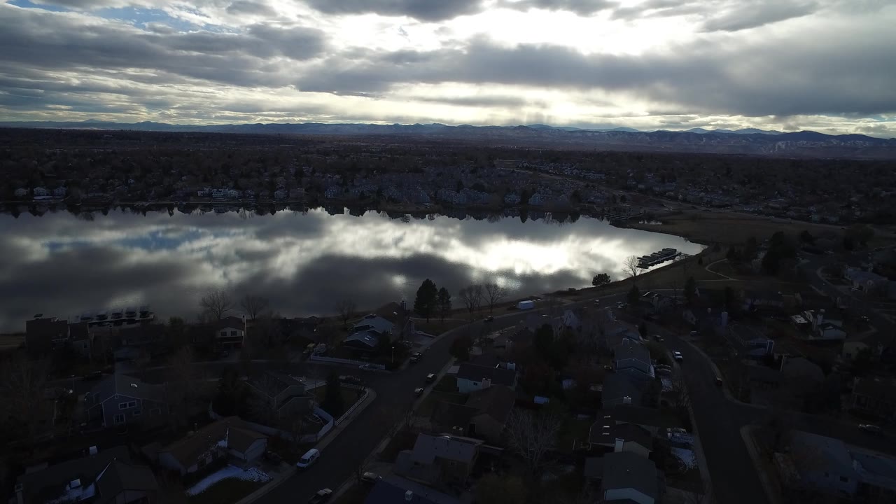 High aerial frame in Westminster, Colorado showing neighborhood rooftops below, the lake reflecting bright clouds across the center, and mountains along the far horizon, with no visible people.