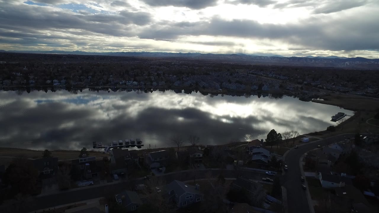 Westminster, Colorado aerial view with bright cloud glare reflected in the lake, houses and trees along the near shore, distant hills, and no visible people.