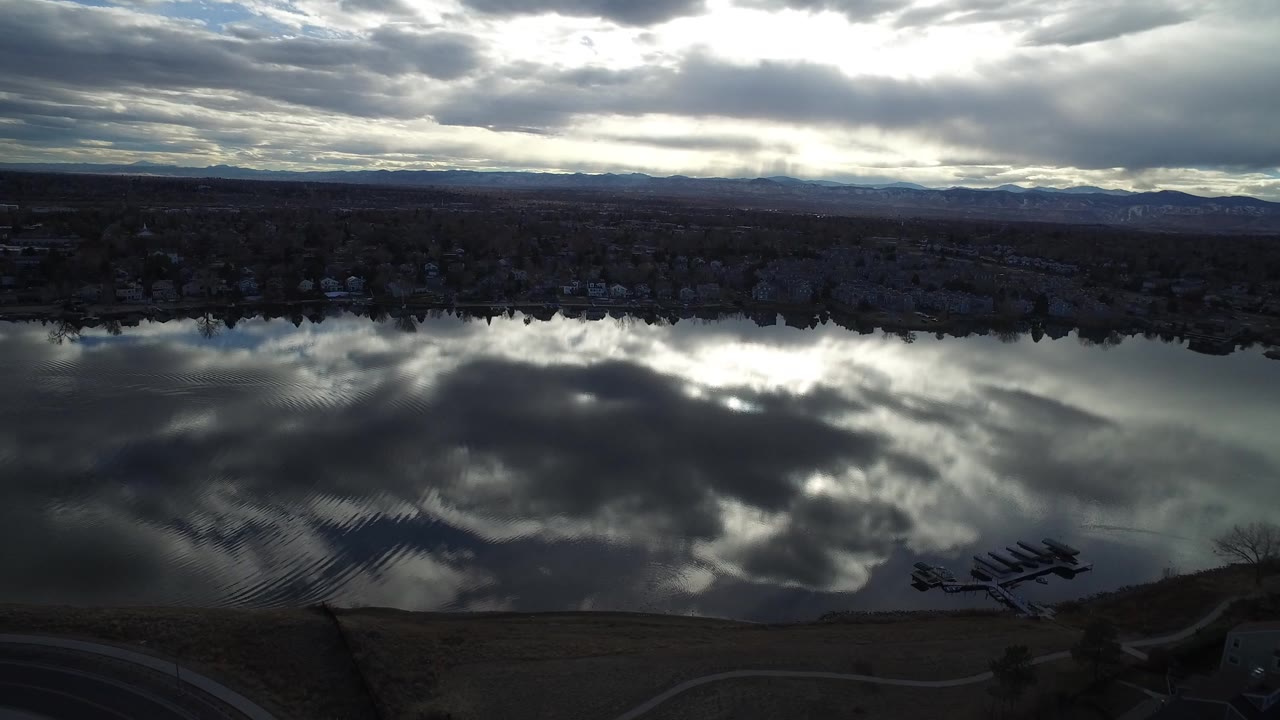 Aerial view in Westminster, Colorado showing dark rippled water reflecting heavy clouds, with a tree-lined shore and distant hills beyond, no visible people.
